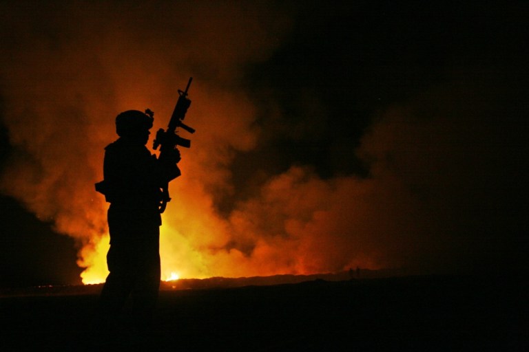 Sgt. Robert B. Brown from Fayetteville, N.C. with Regimental Combat Team 6 watches over the civilian Fire Fighters at the burn pit as smoke and flames rise into the night sky behind him on May 25th, 2007. 