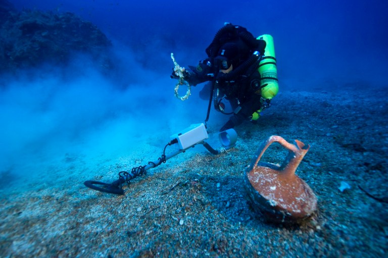 In this undated photo provided by Argo via the Greek Culture Ministry on Thursday, Oct. 9, 2014, a diver with a metal detector holds a copper ship's fitting next to a vase at the site of the Antikythera wreck off the island of Antikythera in southern Greece. The ministry said Thursday that a three-week underwater project to revisit the Roman-era wreck, first investigated more than a century ago, has completed detailed maps of the seabed and pinpointed potential metal artifacts. Divers have recovered a bronze spear that was probably part of a large statue, metal fittings from the ship and a vase. (AP Photo/ARGO via Greek Culture Ministry, Brett Seymour)