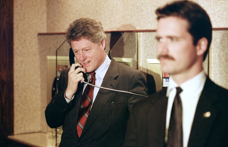 Guarded by a Secret Service agent, Democratic presidential candidate Bill Clinton calls his wife, Hillary, from a pay phone inside his Cleveland hotel after winning primaries in Kentucky and Arkansas, May 26, 1992. (AP Photo/Mark Duncan)