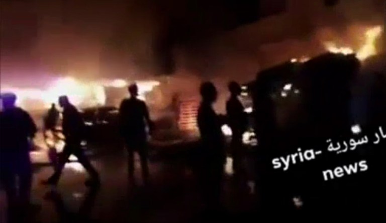 This frame grab from video provided on Wednesday, May, 9, 2018 by Syria News, shows people standing in front of flames rising after an attack on an area known to have numerous Syrian army military bases, in Kisweh, south of Damascus, Syria on Tuesday. The Britain-based Syrian Observatory for Human Rights said the missiles targeted depots and rocket launchers that likely belonged to Iran's elite Revolutionary Guard in Kisweh, killing nine people. (Syria News, via AP)