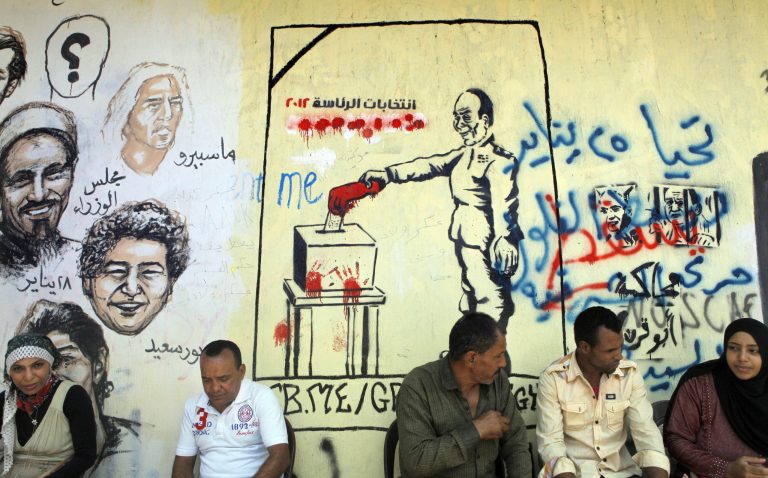   Egyptians sit in front of graffiti showing faces of people killed in the 2011 revolution, right, and the head of the Supreme Council of the Armed Forces, Field Marshal Hussein Tantawi, casting his vote, center, in Tahrir Square in Cairo, Egypt, Friday, June 15, 2012. Judges appointed by Hosni Mubarak dissolved the Islamist-dominated parliament Thursday and ruled that Mubarak's former prime minister can stand in the presidential runoff this weekend, setting the stage for the military and remnants of the old regime to stay in power. Arabic reads 