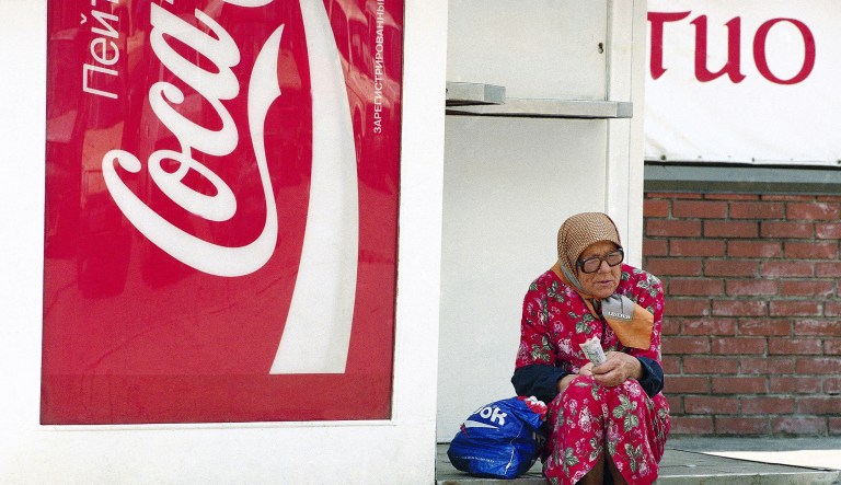 An elderly Russian woman, surrounded by Western symbols of capitalism like the Coca-Cola sign and her plastic Reebok shopping bag, sits along a central Moscow sidewalk and begs for money, June 9, 1995.