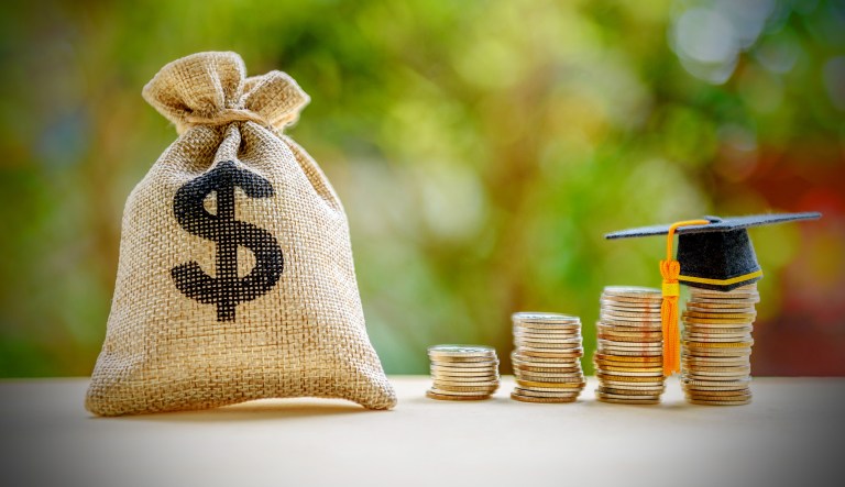 A concept photo of student debt is shown via a dollar bag and a graduation cap on a row of coins on a table.