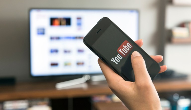 A person uses the YouTube app on a phone while looking at a monitor.