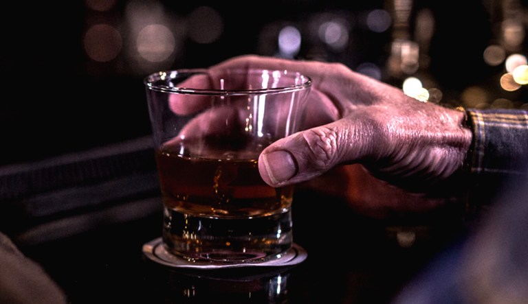 The hand of an old man cups around a glass of whiskey in a barely lit bar.