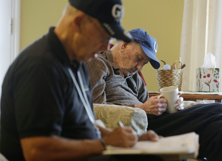 This Nov. 27, 2013, photo, shows caregiver Warren Manchess recording information as Paul Gregoline sits in his favorite chair, in Noblesville, Ind. Home care agencies have shown a willingness to hire older people new to the field who have found a tough job market as they try to supplement their retirement income.(AP Photo/Darron Cummings)