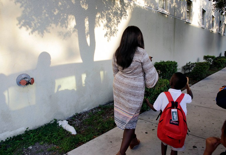 Nadege Cius, left, waits with her son David, 6, for the school bus on the first day of school outside of the Chapman Partnership Homeless Assistance Center, Monday, Aug. 24, 2015, in Miami. Cius lives at the homeless center with her husband and four children. The children got free backpacks, uniforms and school supplies at a giveaway last week. (AP Photo/Lynne Sladky)