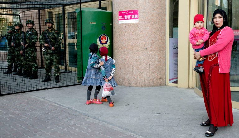 An Uyghur woman carries a toddler as children play near a cage protecting heavily armed Chinese paramilitary policemen on duty in Urumqi in northwest China's Xinjiang Uygur Autonomous Region.
