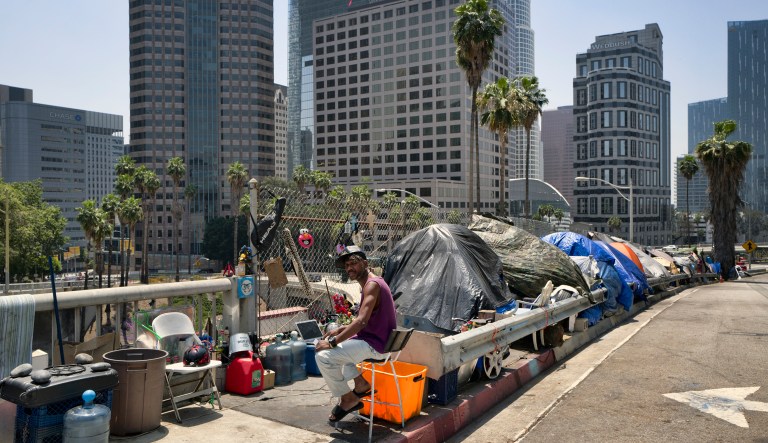 A homeless man sits at his street-side tent by the Interstate 110 freeway along the downtown Los Angeles skyline.