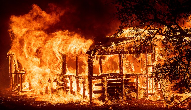 FILE - In this Thursday, July 26, 2018 file photo, a structure burns as the Carr fire races along Highway 299 near Redding, Calif.  In the last year, fires have devastated neighborhoods in the Northern California wine country city of Santa Rosa, the Southern California beach city of Ventura and, now, the inland city of Redding. Hotter weather from changing climates is drying out vegetation, creating more intense fires that spread quickly from rural areas to city subdivisions, climate and fire experts say. But they also blame cities for expanding into previously undeveloped areas susceptible to fire. 