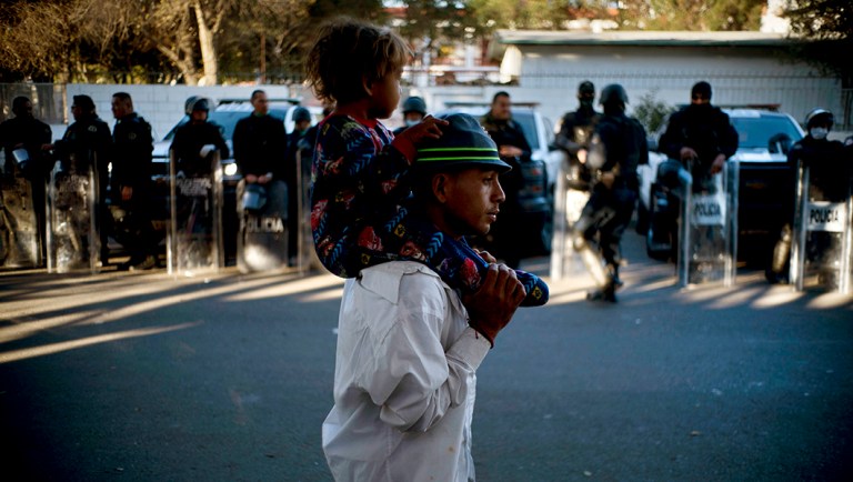 A migrant carries a child past Mexican police who stand guard outside the Benito Juarez Sports Center.
