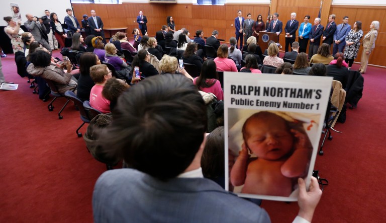 A demonstrator holds a sign as Gov. Ralph Northam, at podium, gives a news conference at the Capitol in Richmond, Va., Thursday, Jan. 31, 2019. Northam made a statement and answered questions about the late term abortion bill that was killed in committee.
