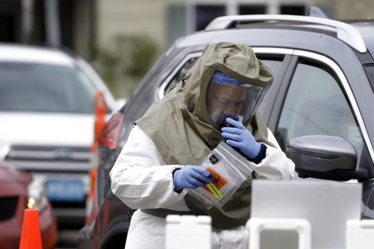 A medical worker wearing protective equipment carries packets while administering tests for the coronavirus, Tuesday, April 28, 2020, at a testing site in a parking lot of a hospital, in Somerville, Mass.