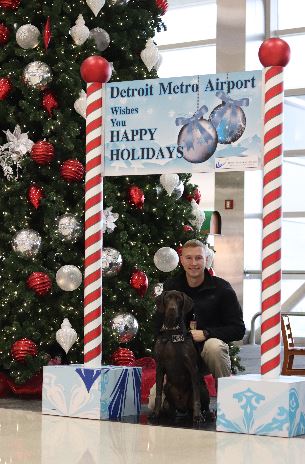 Bobi (left) is pictured near a person, a holiday sign, and a Christmas tree at Detroit Metropolitan Airport.