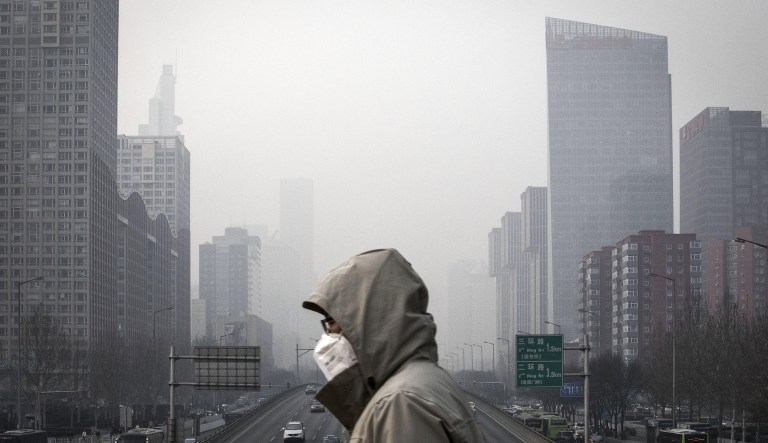 A man wearing a face mask walks on a footbridge as buildings shrouded in haze stands in the background in Beijing, China, on Friday, Jan. 6, 2017.