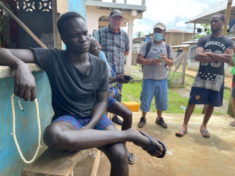 A migrant sits on a bench in Panama after exiting Darien Gap. Rep. Tom Tiffany, a Wisconsin Republican (in plaid shirt) looks on.