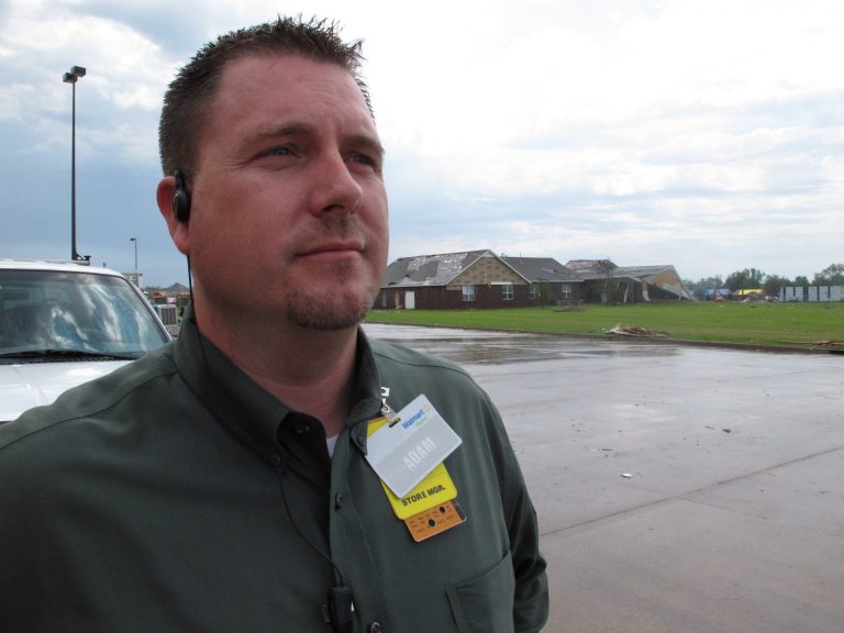 In this Thursday, May 23, 2013 photo, Wal-Mart manager Adam Stutzman stands behind his store in Moore, Okla. More than five dozen workers, customers and passersby took shelter in the store's meat room from Monday's tornado, which collapsed the church in the background. (AP Photo/Allen Breed)
