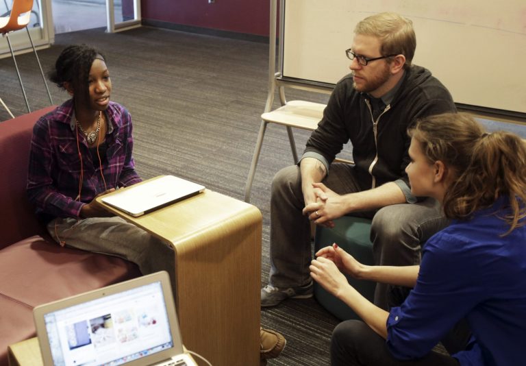 English teacher Tom Rademacher talks with his high school juniors Kierra Murray, left, and Ana Silverman, right, Tuesday, Dec. 2, 2014, at Fair School in Minneapolis. (AP Photo/Jim Mone)