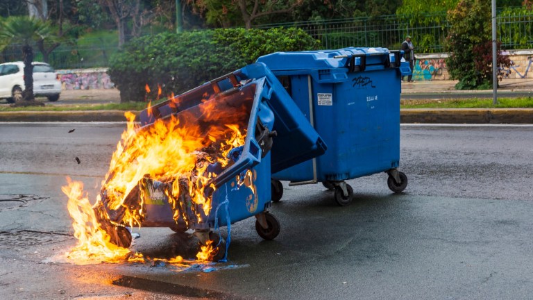 A plastic dumpster bin melts as it is enveloped in flames. Another plastic bin sits beside it.