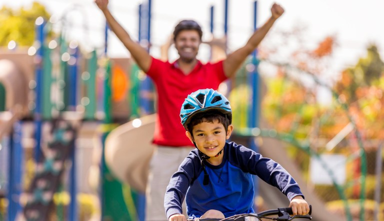 A father cheers on as his son rides a bicycle.