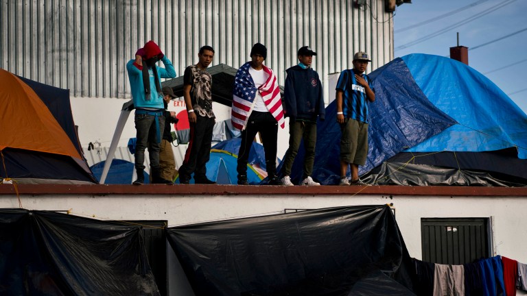 Migrants, one wearing a U.S. flag, stands on a roof where they installed their tents at a new shelter where they were transferred to after the sanitary conditions worsened at a previous shelter in Tijuana, Mexico. (AP Photo/Ramon Espinosa)