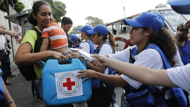 A woman with a child receives an empty container for water and water purification pills during the Red Cross' first aid shipment in Caracas, Venezuela, Tuesday, April 16, 2019. In late March, the Red Cross federation announced it would soon begin delivering assistance to an estimated 650,000 people and vowed that it would not accept interference from either side of the polarized country. 