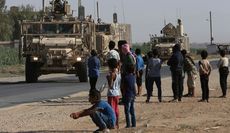 FILE - In this July 26, 2017 file photo, Syrians look at a U.S. armored vehicle convoy on a road that leads to Raqqa, northeast Syria. Amnesty International, in a report Tuesday, June 5, 2018, accused the U.S. and its allies of showing little regard for civilians' lives while attacking the Syrian city that was once the self-styled capital of the Islamic State group. The report said that the U.S.-led coalition's 2017 assault on Raqqa killed hundreds of civilians and reduced sections of the city to rubble. A coalition spokesman calls the allegations "absurd and grossly inaccurate."