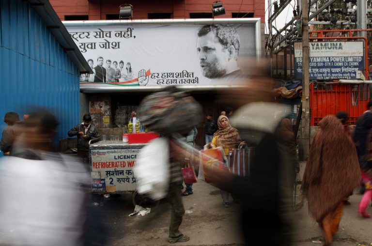 In this Feb. 8, 2014 photo, Indian people walk past a Congress party hoarding displaying portrait of their vice president Rahul Gandhi, right, and party symbol in old quarters of New Delhi, India. Election season in India always comes with heaps of promises, as political parties try to woo voters with everything from subsidized fuel and electricity to free laptops, spice grinders, even goats and cows.  But there are growing concerns that freebies will hurt the country's sputtering economy. (AP Photo/Tsering Topgyal)