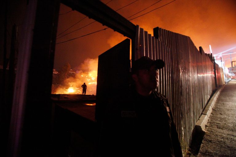 A man stands behind a wall as an out of control forest fire destroys homes in the city of Valparaiso, Chile, Sunday April 13, 2014. Firefighters struggled for a second night to contain blazes that reached this port city, killing at least a dozen people, destroyed hundreds homes and has forced the evacuation of thousands. (AP Photo/Luis Hidalgo)