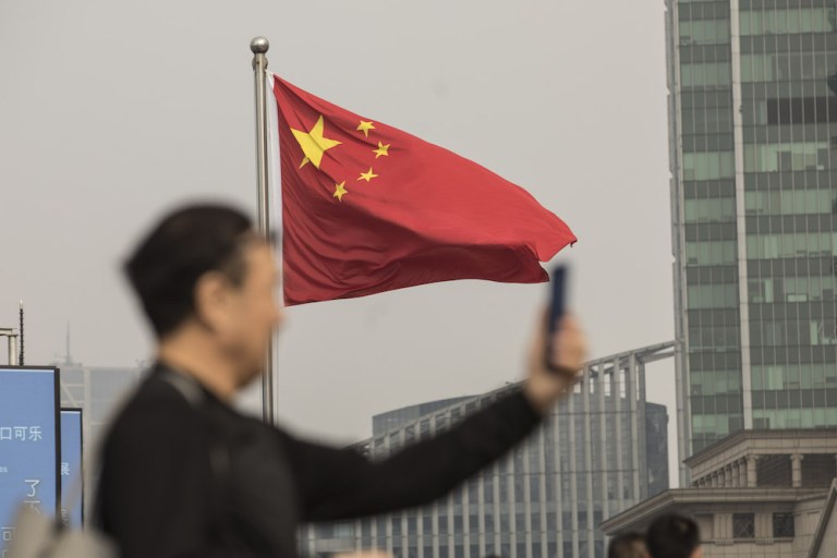 A Chinese national flag flies as a man uses a smartphone in Shanghai, China, on Tuesday, Nov. 27, 2018. Apple, which has lost a fifth of its value in a tech market rout since October, is poised for another setback after U.S. PresidentÂ Donald TrumpÂ suggested that 10 percent tariffs could be placed on mobile phones, like the iPhone, and laptops made in China. 