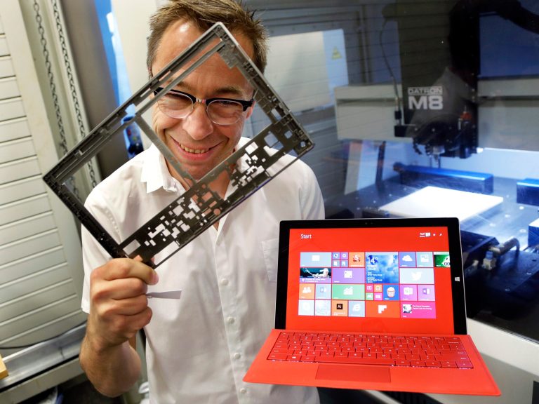 In this photo taken July 3, 2014, Ralf Groene, chief designer for the Microsoft Surface tablet computer, poses for a photo with a Surface 3 Pro tablet, right, in front of a milling machine used to produce parts for the computer, like the one he is holding at left, in Redmond, Wash. As Microsoft competes with Apple and other companies for hardware sales, the software giant has put a new emphasis on design. (AP Photo/Ted S. Warren)