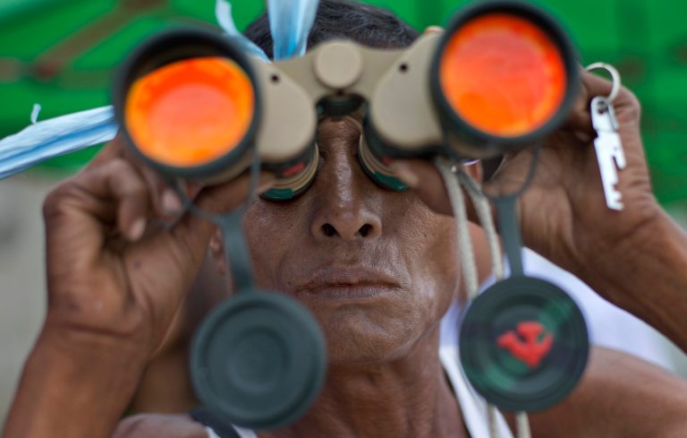 In this Tuesday, Aug. 19, 2014 photo, a man uses a pair of binoculars to get a glimpse of recovery efforts of an ancient bell in the Yangon River in Yangon, Myanmar. Unsubstantiated rumors that the 270-ton mysterious bell has been spotted have sent thousands of curious spectators flocking to the banks of the river. The world's largest copper bell, believed to have been lying deep beneath the riverbed for more than four centuries. (AP Photo/Gemunu Amarasinghe)