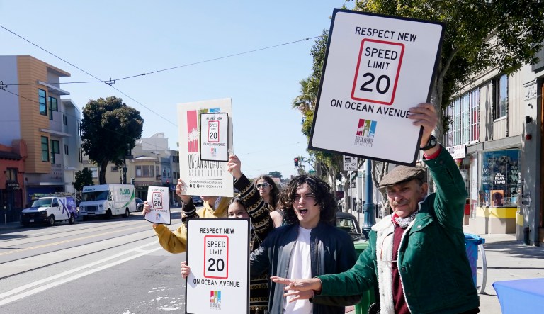 Ocean Avenue Association executive director Pierre Smit, right, and Lick-Wilmerding High School sophomores hold up signs for oncoming traffic at a Slow Down on Ocean Ave event in San Francisco, Wednesday, March 9, 2022. (AP Photo/Jeff Chiu)