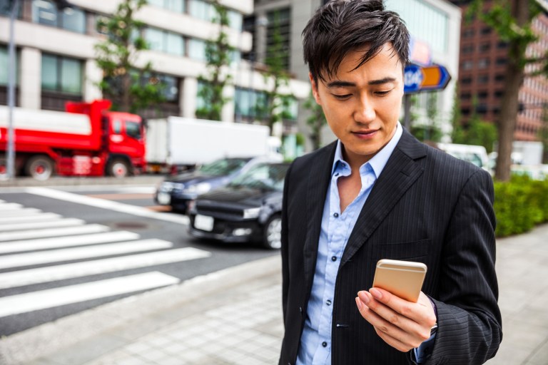 Man in business outside the office building in downtown Tokyo financial district waiting for a meeting in Tokyo, Japan. He's walking with suit and suitcase to the office. Waiting colleagues and having phone call and text messages on the sidewalk, while looking for car pooling.