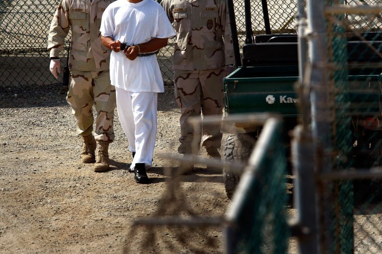 U.S. military guards walk a detainee through Camp Delta in the Guantanamo Bay detention center on March 29, 2010 in Guantanamo Bay, Cuba. (Photo by John Moore/Getty Images)
