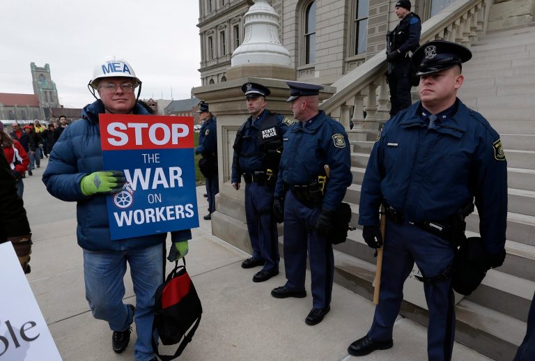 A protester, wearing a Michigan Education Association helmet, walks past Michigan State Police at the State Capitol in Lansing, Mich., in 2012. (AP Photo/Paul Sancya)