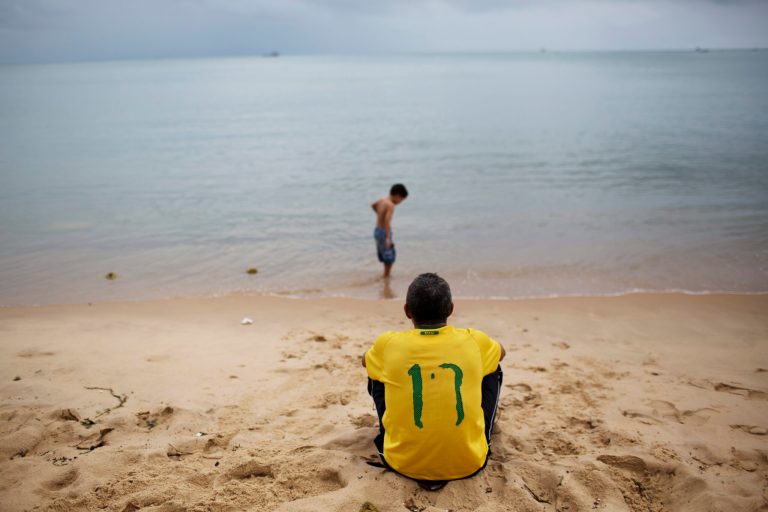 Paulo Cesar Da Silva, wearing his Brazilian soccer T-shirt with a weathered #10, the number associated with soccer star Neymar, looks out to the ocean as his son Gabriel wades along the shoreline in Fortaleza, Brazil, Thursday, July 3, 2014. For two whole days now, the globe has gone without World Cup soccer matches in this most electrifying of tournaments in decades. There were no matches on Wednesday and Thursday as teams still standing took a break before heading into Friday's quarterfinal matches. (AP Photo/Rodrigo Abd)