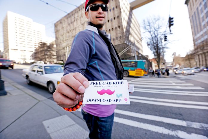 In this photo taken Friday, March 14, 2014, Jerad Bernard hands out cards to passersby offering one free ride through the Lyft ridesharing service in Seattle. In a fight pitting upstart technology and traditional business, app-based car share companies and traditional taxis are fighting for supremacy. (AP Photo/Elaine Thompson)