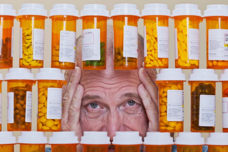 Senior man with an unhappy, depressed expression and his hands framing his face looking through an opening in rows of prescription medication. Health care and medication cost can overwhelm many senior citizens.