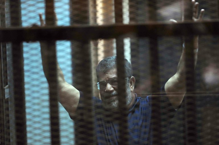 Ousted Egyptian President Mohammed Morsi raises his hands as he sits behind glass in a courtroom, in a converted lecture hall in the national police academy in an eastern Cairo suburb, Egypt, Saturday, May 16, 2015. (AP Photo/Ahmed Omar)
