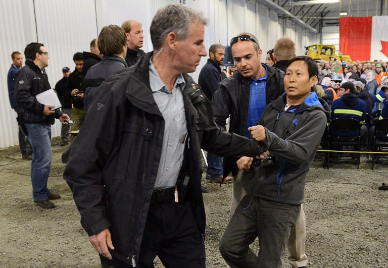 In this Friday, Aug. 23, 2013 photo, Li Xue Jiang, of the People's Daily, China's largest newspaper, is hauled to the back of the room by Royal Canadian Mounted Police as Canadian Prime Minister Stephen Harper answers questions while visiting Xstrata Nickel's Raglan Mine in the northern Nunavik region of Quebec. When staff did not recognize Li's request to answer a question he tried to take the microphone and a tussle erupted. Canada said Friday, Aug. 22, 2014, 