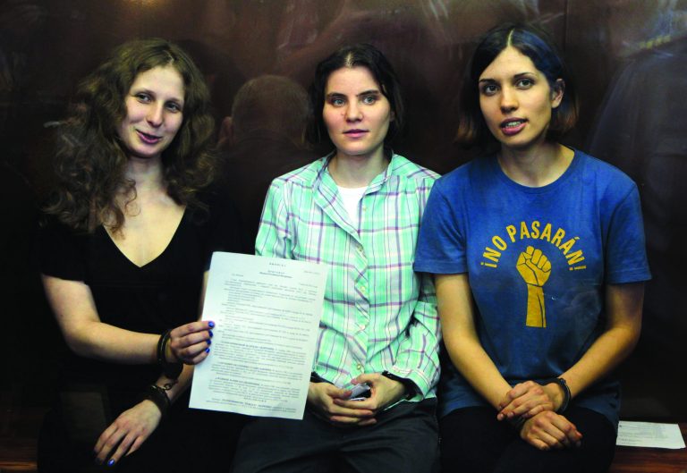 FILE - In this Friday, Aug. 17, 2012 file photo, feminist punk group Pussy Riot members, from left, Maria Alekhina, Yekaterina Samutsevich, and Nadezhda Tolokonnikova show the court's verdict as they sit in a glass cage at a courtroom in Moscow. The two-year prison sentence handed down to Pussy Riot for a provocative protest inside a Moscow cathedral called attention to just how hard President Vladimir Putin is clamping down on minor displays of dissent. But Russia isn't the only country where people are punished for offenses that many in the West might consider trivial. (AP Photo/Mikhail Metzel, File)