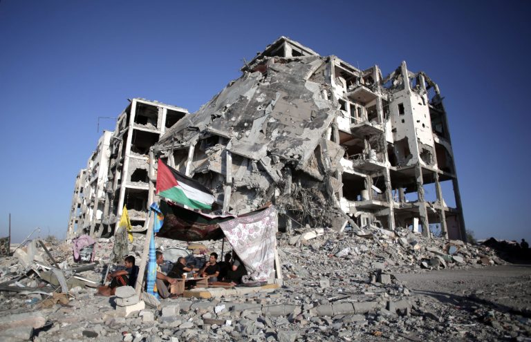 FILE- In this Monday, Aug. 11, 2014 file photo, Palestinian Ziad Rizk, sits with others in a shelter made of a blanket stretched over four boles next to one of the destroyed al-Nada Towers, where he lost his apartment and clothes shop, in the town of Beit Lahiya, northern Gaza Strip. Dozens of residents the al-Nada Towers took advantage of the latest truce to visit their homes, salvage what they can and exchange tips on how best to seek and secure compensation for their homes. (AP Photo/Khalil Hamra, File)