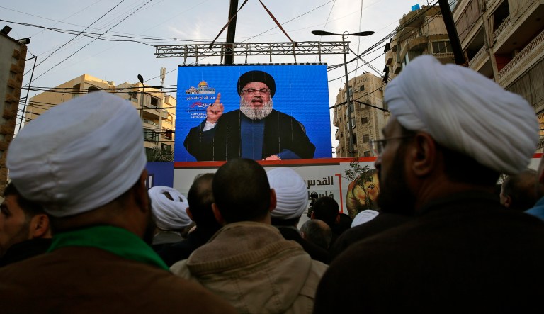 In this photo from Monday, people listen to Hezbollah leader Sheikh Hassan Nasrallah as he speaks on a screen via a video link. On Thursday, the business committee of the British Parliament will debate whether to proscribe Hezbollah's political wing as a terrorist organization. (AP Photo/Bilal Hussein)