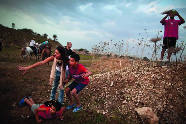 Israelis take cover as an air raid siren warns of incoming rockets from Gaza, next to an Iron Dome defense system in Tel Aviv, Saturday, Nov. 17, 2012. Israel bombarded the Hamas-ruled Gaza Strip with nearly 200 airstrikes early Saturday, the military said, widening a blistering assault on Gaza rocket operations to include the prime minister's headquarters, a police compound and a vast network of smuggling tunnels. (AP Photo/Oded Balilty)
