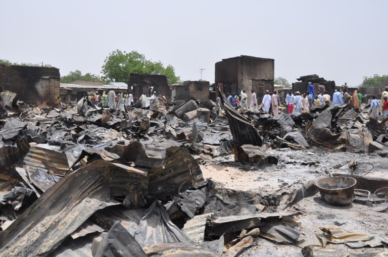 In this photo taken Sunday, May 11, 2014, people stand outside burnt houses following an attack by Islamic militants in Gambaru, Nigeria.  Many brutalized residents of the once bustling town of Gamboru said Monday May 12, 2014, they are moving across the border to Cameroon because they cannot trust the Nigerian government or military to protect them, after repeated attacks by Islamic militants, including an attack a few days ago that killed some hundreds of people with more than 1,000 shops, dozens of homes and 314 trucks and cars bombed and burned out.(AP Photo/Jossy Ola)