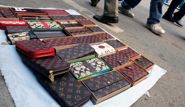 Pedestrians walk past fake Louis Vuitton and Coach brand purses being sold on a street.