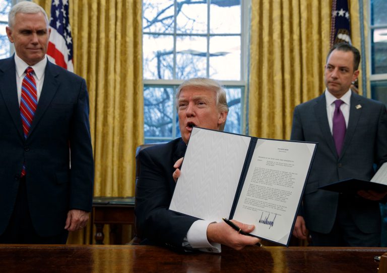 DAY 4 - In this Jan. 23, 2017, file photo, Vice President Mike Pence, left, and White House Chief of Staff Reince Priebus, right, watch as President Donald Trump shows off an executive order to withdraw the U.S. from the 12-nation Trans-Pacific Partnership trade pact agreed to under the Obama administration in the Oval Office of the White House in Washington.