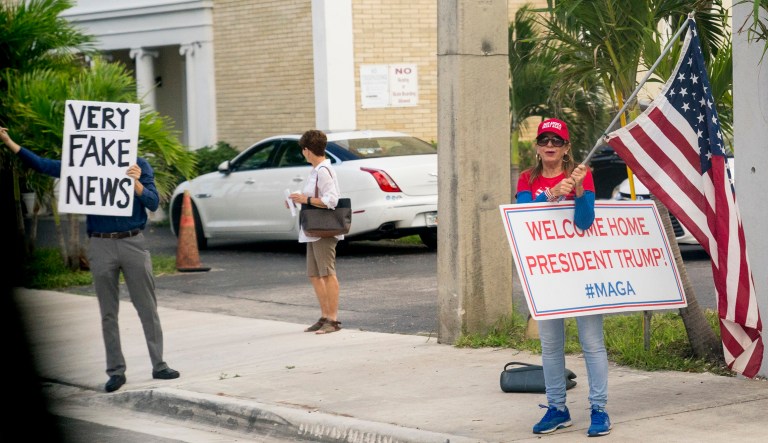 Pedestrians hold up signs that read "Very Fake News" and "Welcome Home President Trump #MAGA" as the motorcade carrying President Trump makes its way . 