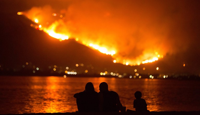 A family sits along the shore of Lake Elsinore as they watch the Holy Fire burn in the distance on Thursday night, Aug. 9, 2018 in Lake Elsinore, Calif.  More than a thousand firefighters battled to keep a raging Southern California forest fire from reaching foothill neighborhoods Friday before the expected return of blustery winds that drove the flames to new ferocity a day earlier. 